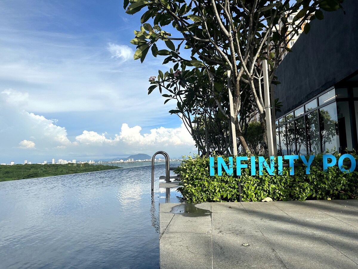 An infinity pool stretches along the edge, reflecting the skyline above. Lush greenery is seen along the poolside, framing the calm water. The backdrop features a clear blue sky with clouds, creating a serene environment.