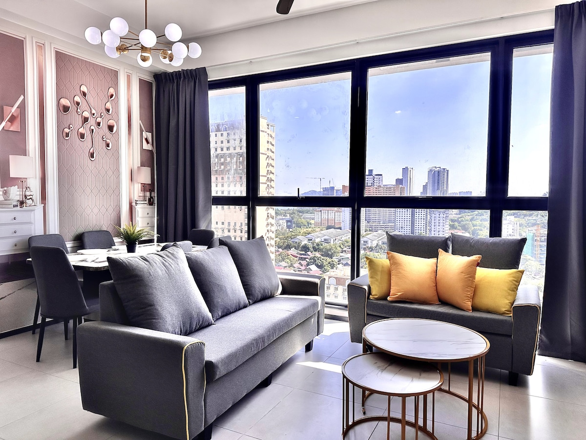 A modern living area features large floor-to-ceiling windows that offer a panoramic view of the city skyline. A grey sofa and armchair are complemented by vibrant orange throw pillows. Two round coffee tables sit in front of the seating area, and a stylish light fixture hangs overhead.