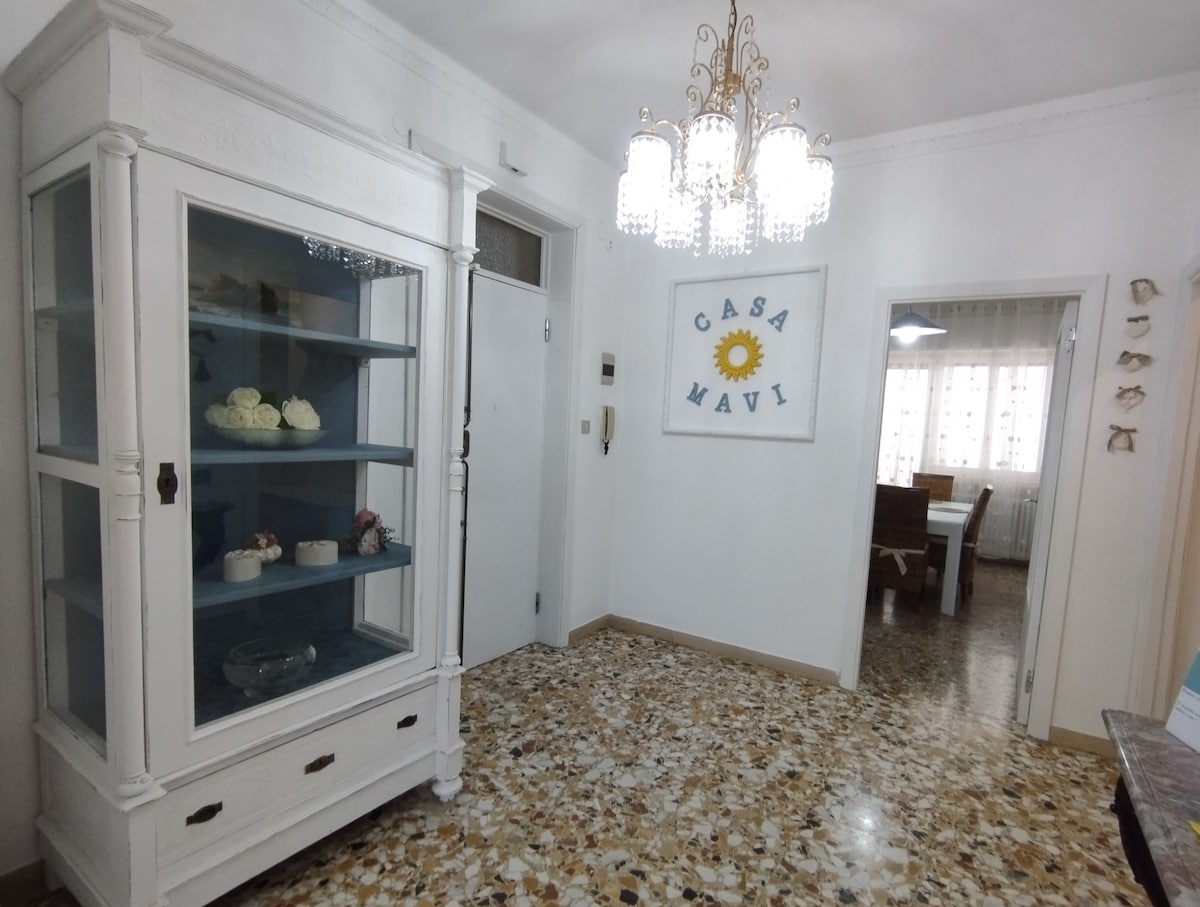 An entrance hall is highlighted by a white cabinet with glass doors, showcasing decorative items. A chandelier with multiple crystals hangs from the ceiling, illuminating the space. The floor features a mosaic pattern, while a sunburst wall decoration with the name 'Casa Mavi' adds a welcoming touch.