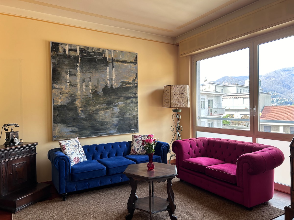A cozy seating area features a blue tufted sofa and a pink tufted couch, accompanied by a wooden coffee table. A large artwork adorns the wall, and a lamp stands beside the window, providing a view of the surrounding mountains.