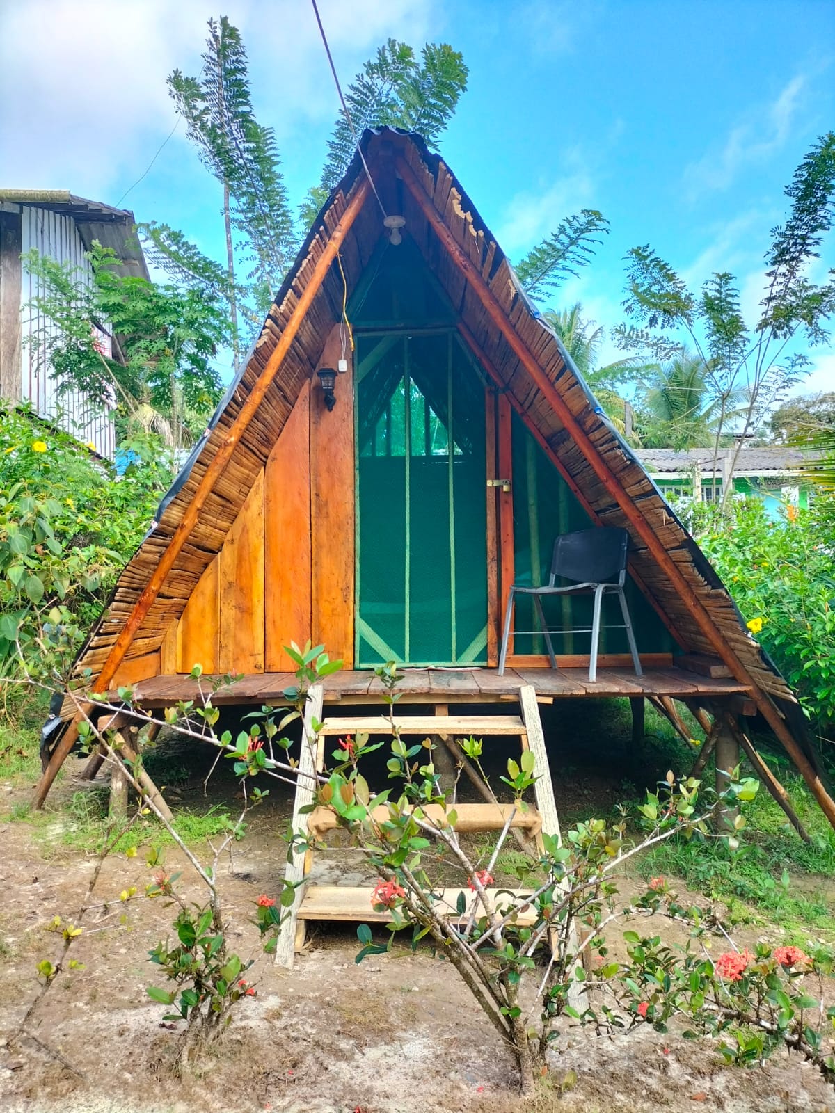 A charming triangular cabin is nestled amidst lush greenery, featuring wooden exterior walls and a green mesh door for ventilation. A simple chair is placed on a small porch steps leading to the entrance, surrounded by flowering plants and shrubs.