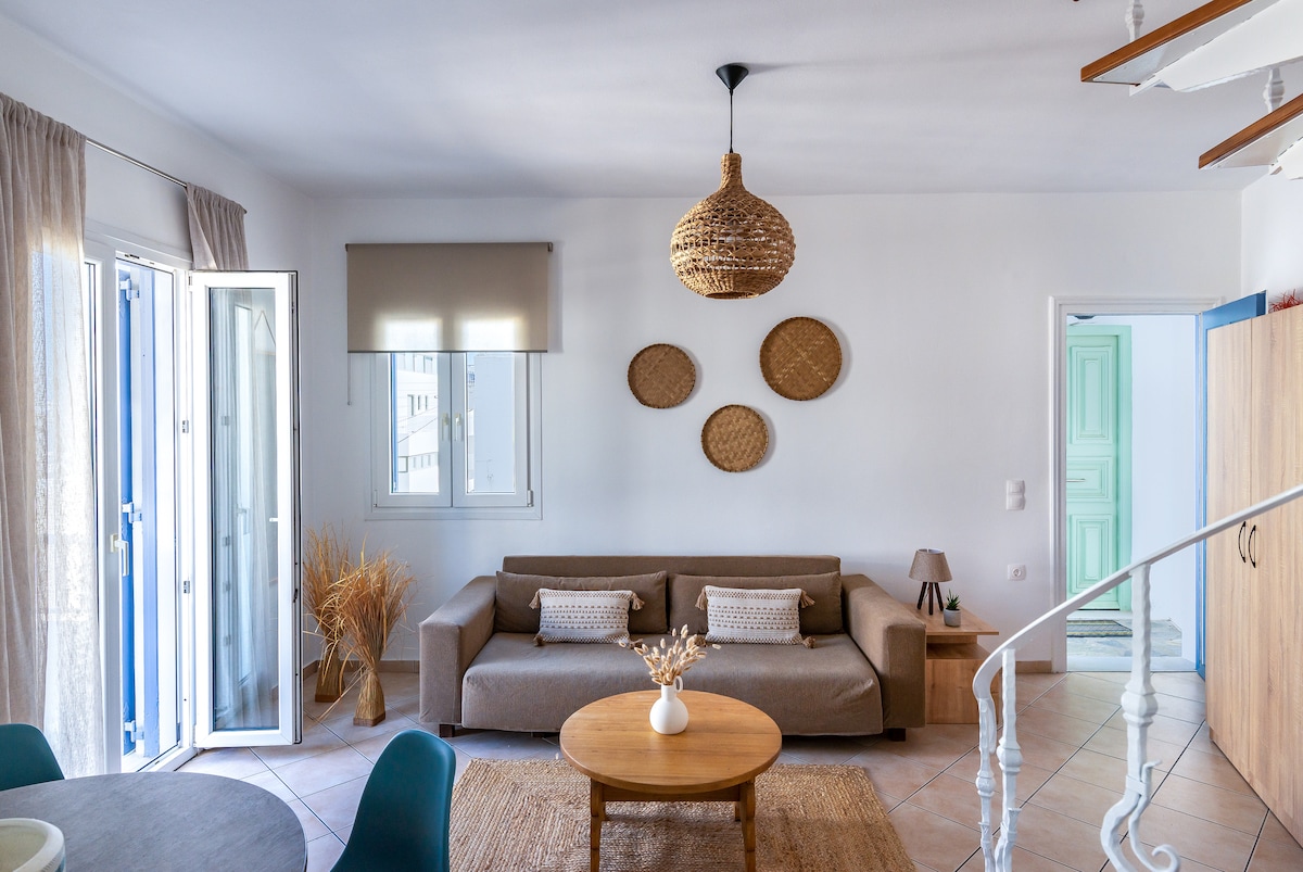A living area is presented with a light-colored sofa and a round wooden coffee table. Natural woven decor adorns the walls. Two doors provide access to the outdoors, allowing for ample natural light. The flooring features tiles in a neutral tone, contributing to the cozy ambiance.