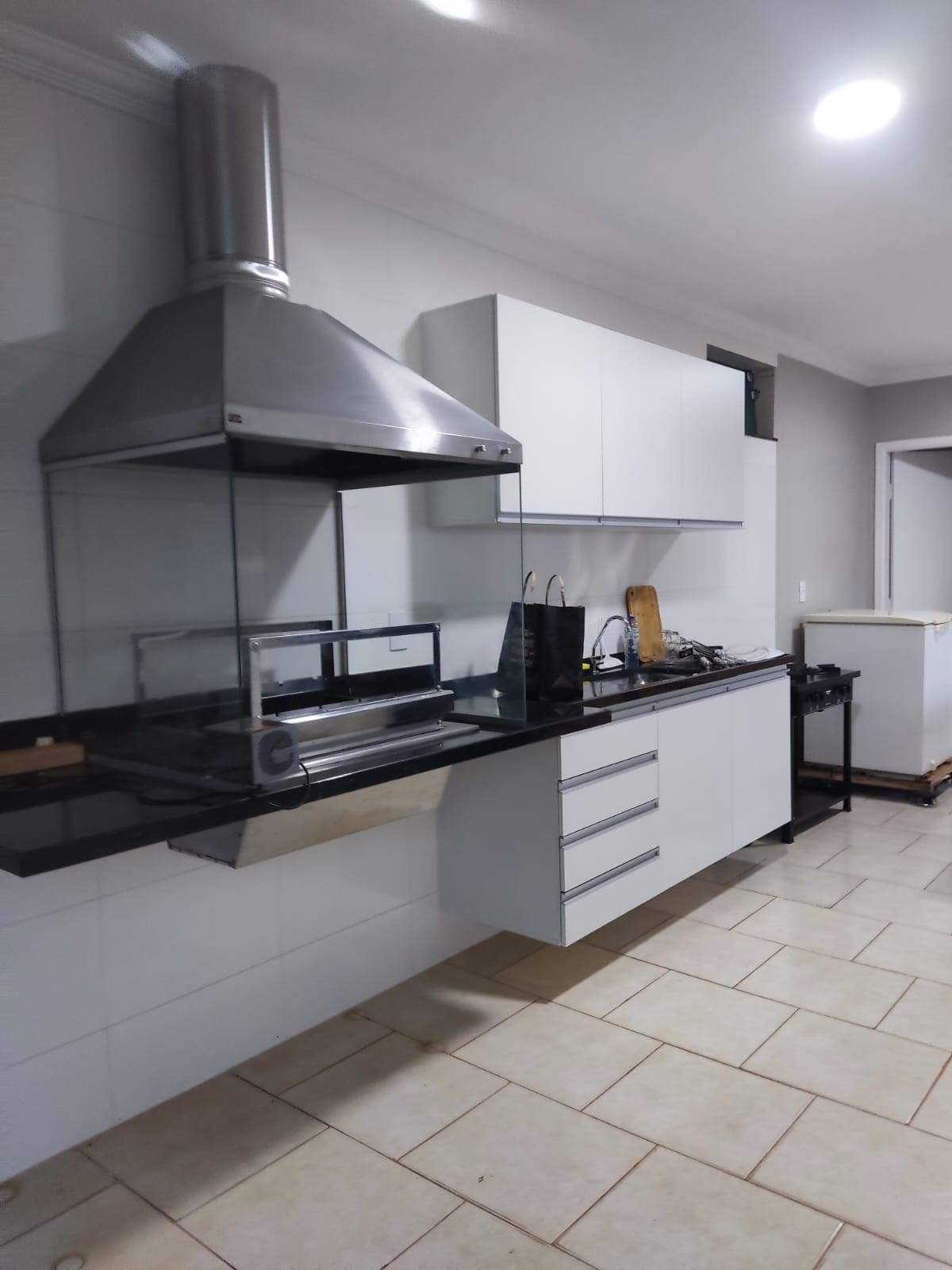 A modern kitchen features a sleek black countertop with white cabinetry. A stainless-steel hood is positioned above a gas stove, while a glass partition adds a contemporary feel. Natural light illuminates the area, highlighting the organized layout and functional appliances.