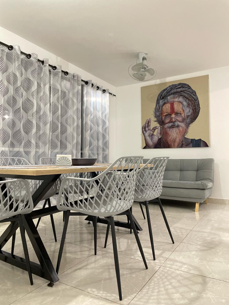 A dining area features a rectangular table surrounded by six woven chairs. A modern sofa provides additional seating. Decorative sheer curtains frame the window, and an artwork piece adorns the wall, creating an open and inviting space.