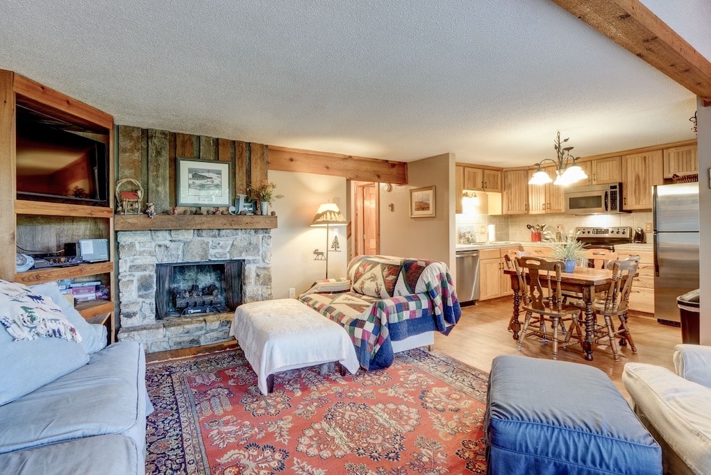 A living area features a stone fireplace, surrounded by comfortable seating arrangements, including a patterned sofa and armchairs. A wooden dining table with multiple chairs is positioned nearby, with a well-equipped kitchen visible in the background, creating a cozy family space.