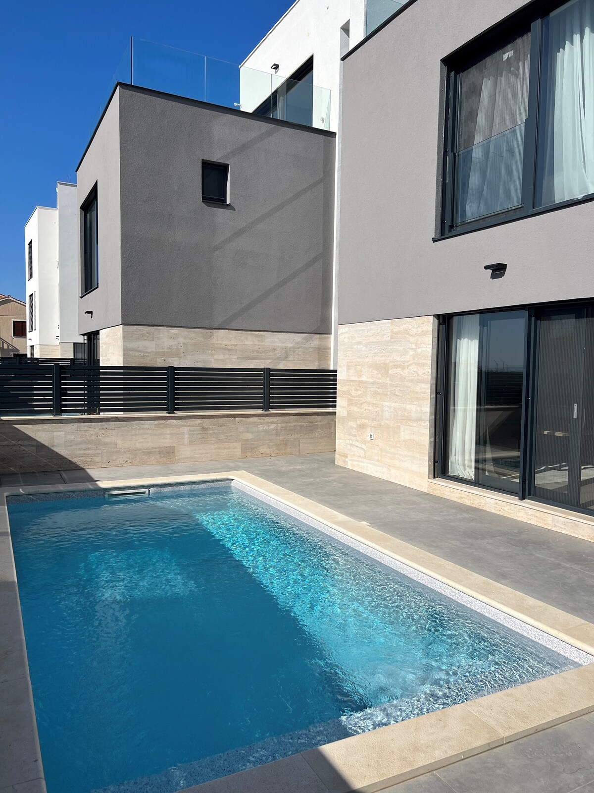 A modern pool area is depicted, featuring a private heated pool with clear blue water. Surrounding walls are composed of sleek materials, and tall glass doors provide access to the interior. The bright sky and sunlight enhance the spacious outdoor atmosphere.