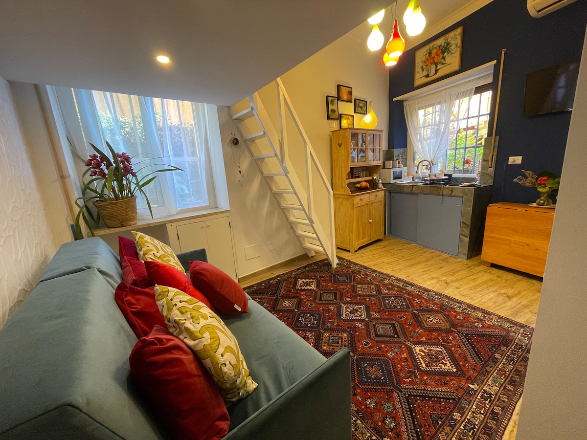 A cozy living area features a comfortable sofa adorned with colorful pillows, positioned near a large window. A small staircase leads to a loft area. The kitchenette, equipped with cabinets and essential appliances, complements the inviting atmosphere created by warm lighting.