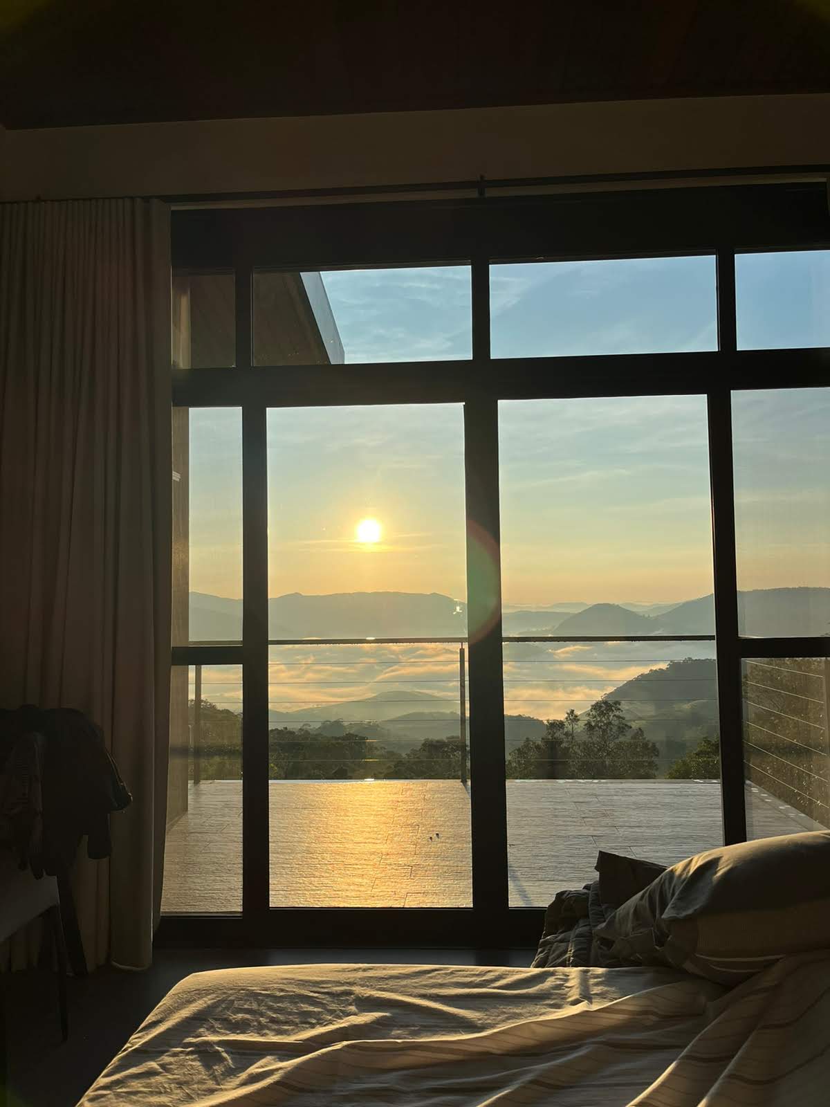 A spacious room is illuminated by morning light, with large glass windows offering views of a serene sunrise over misty mountains. A comfortable bed is positioned near the window, providing a peaceful setting to enjoy the natural scenery.