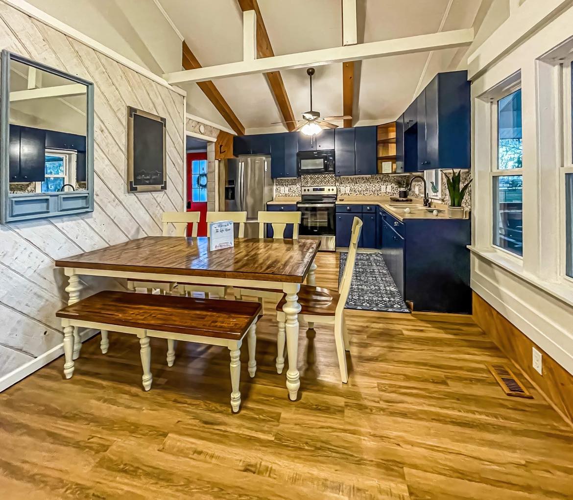 A dining area is featured, showcasing a large wooden table with a matching bench and chairs. Blue cabinetry is visible in the background, complemented by stainless steel appliances. Natural light enters through multiple windows, enhancing the inviting atmosphere of the space.