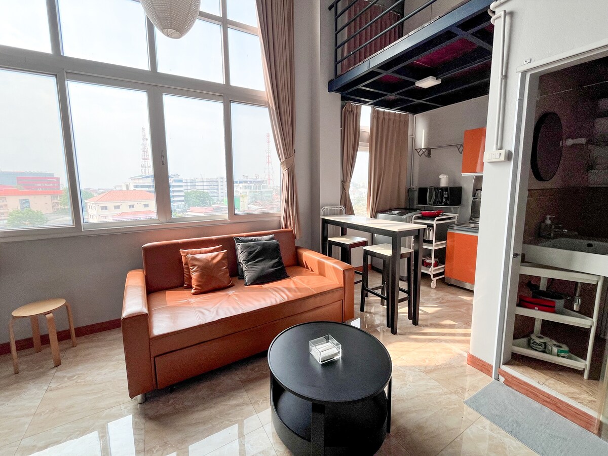 A modern living area features a leather sofa with colorful cushions, a round coffee table, and a compact dining table. Large windows allow natural light to fill the space, offering views of the surrounding city. The compact kitchen is visible in the background.