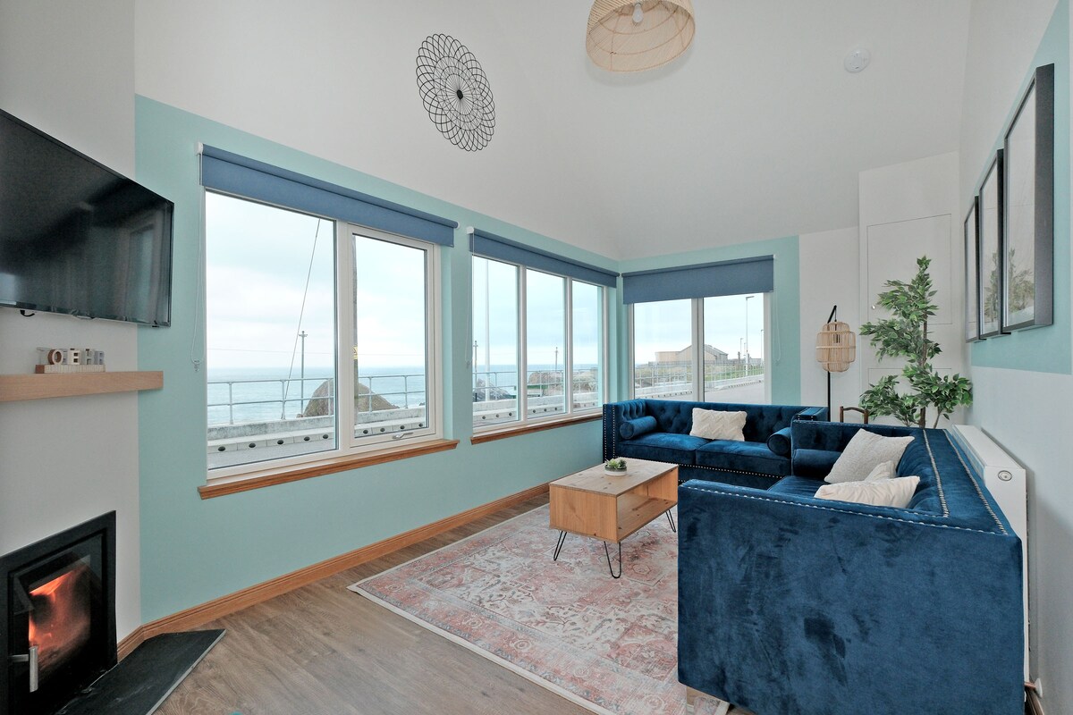 A spacious living area features large windows offering views of the sea. A cozy blue sofa and a light wooden coffee table complement the room. Soft lighting is provided by a modern fixture above, while a potted plant adds a touch of greenery.