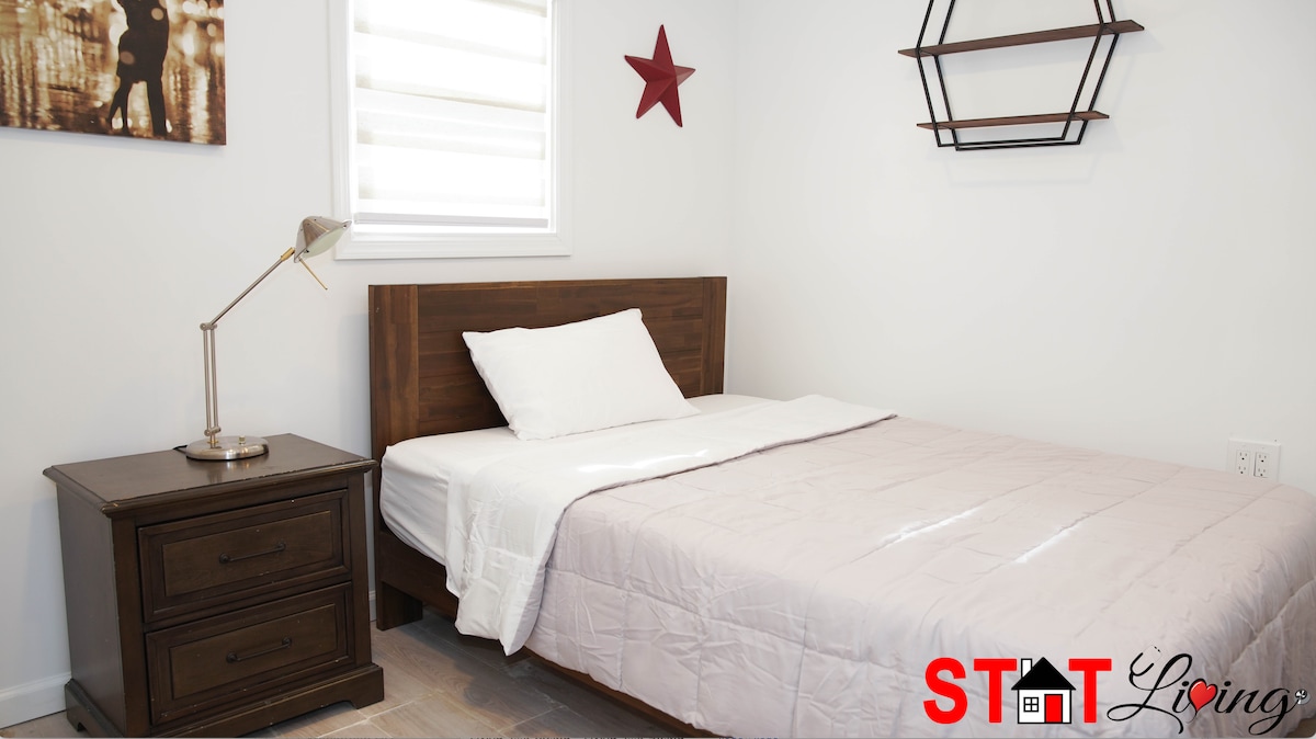 A tidy sleeping area features a wooden bed frame with a light-colored comforter. A bedside table with drawers accompanies the bed, and a modern lamp provides soft lighting. A simple wall shelf and a decorative star enhance the room's minimal aesthetic.