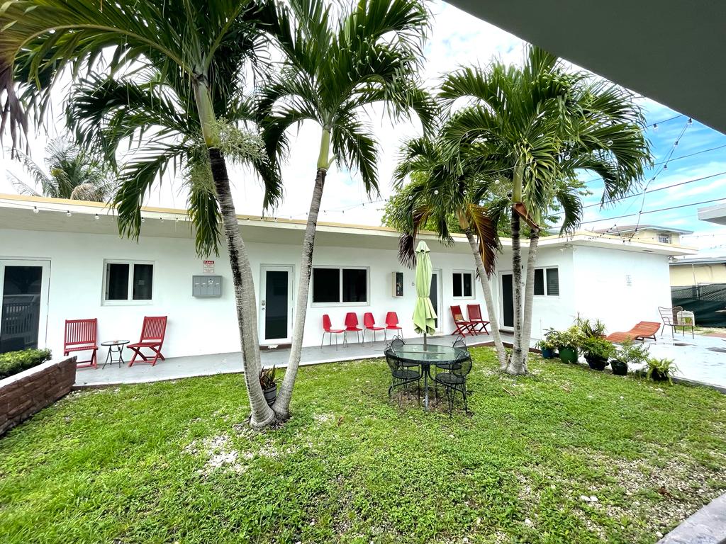 A landscaped outdoor area features a green lawn surrounded by palms. A round table, accompanied by several chairs, is positioned at the center. Several red chairs are placed along the building, with windows visible in the background.