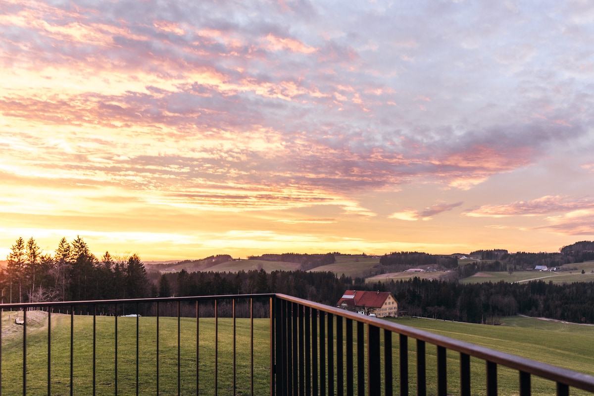 A balcony railing frames a stunning sunset view, with vibrant hues of orange and pink in the sky above rolling hills and fields. Distant trees and a farmhouse are visible, blending naturally into the serene landscape.