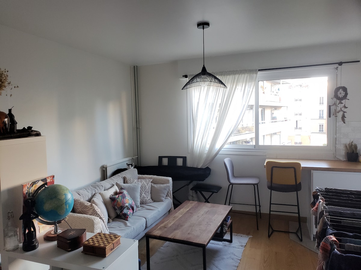 A bright living area is furnished with a comfortable couch adorned with various cushions. A wooden coffee table sits in the center, while a window allows natural light to fill the space. Additional seating options include a stool and chairs near a workspace.