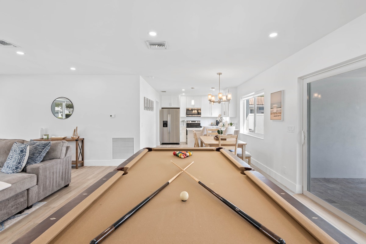 A spacious living area features a billiard table in the foreground, with cues resting on the felt. In the background, a modern kitchen can be seen, highlighted by stylish pendant lighting and a dining area. Soft neutral tones create a welcoming ambiance.