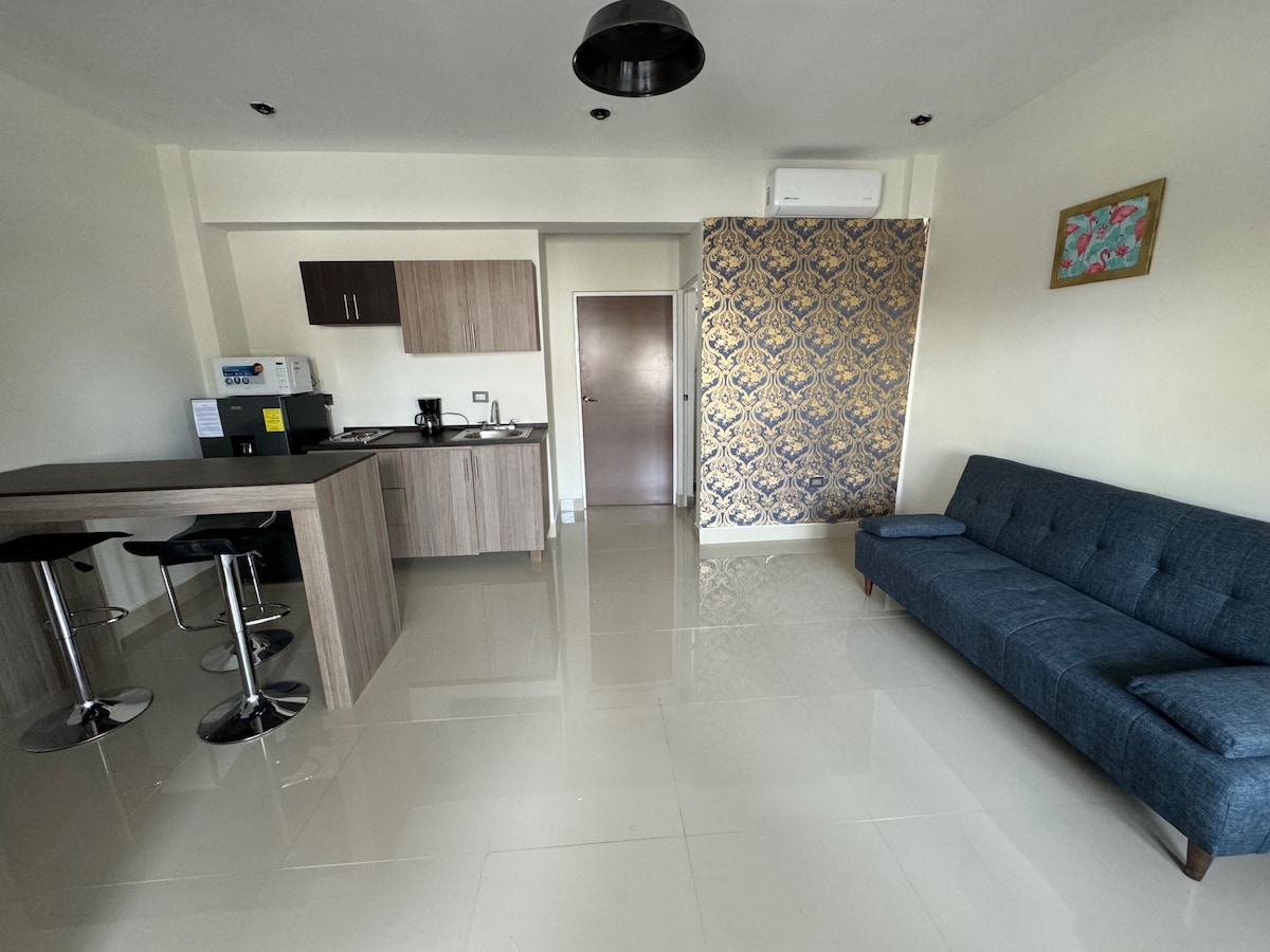 A modern living space features a cozy blue sofa and a simple dining area with high stools. A kitchenette with wooden cabinetry and essential appliances is visible. The walls are adorned with a decorative piece, and natural light brightens the polished floor.