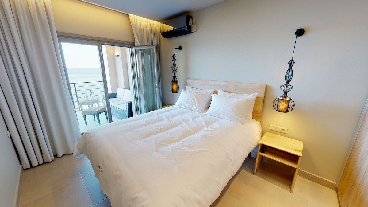 A serene bedroom is furnished with a spacious bed and minimalist nightstand. Natural light enters through glass doors leading to a small balcony, which offers a view of the surroundings. Soft curtains frame the window, complemented by stylish pendant lights on each side of the bed.