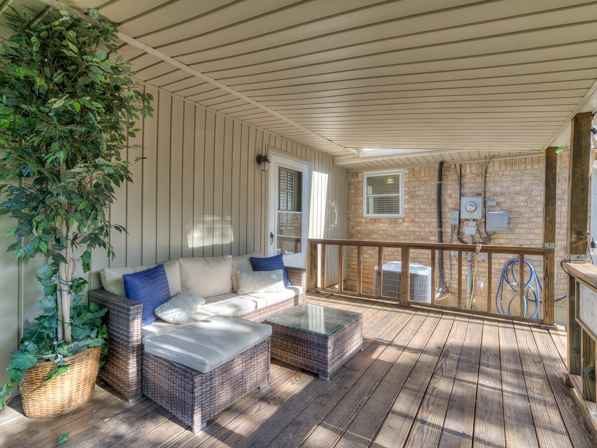 An outdoor seating area features a comfortable couch with blue accent pillows and a glass-topped coffee table. Lush greenery is present in a large planter, while the surrounding wood deck provides ample space for relaxation.