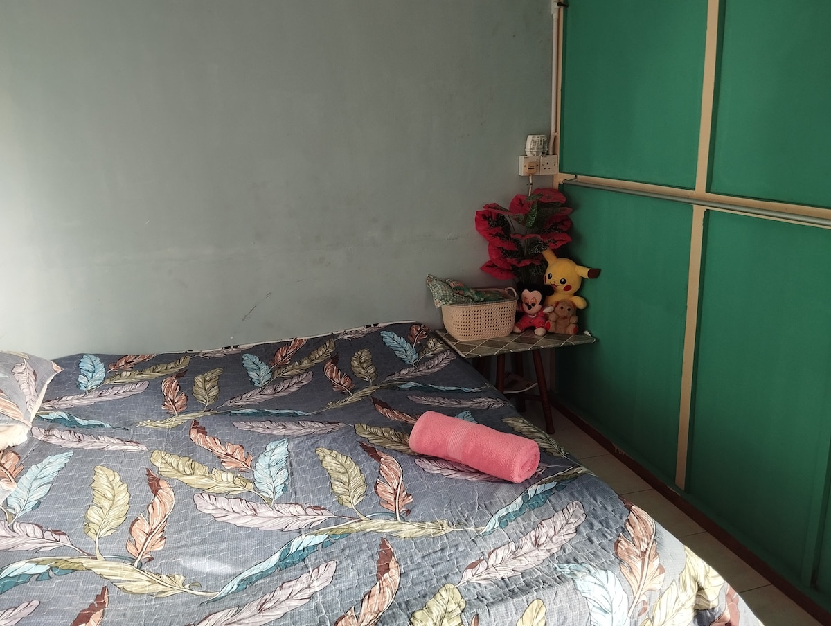A cozy bedroom features a bed adorned with a colorful leaf-patterned quilt. A small table holds a basket with plush toys and a rolled towel, while green walls create a calming backdrop.
