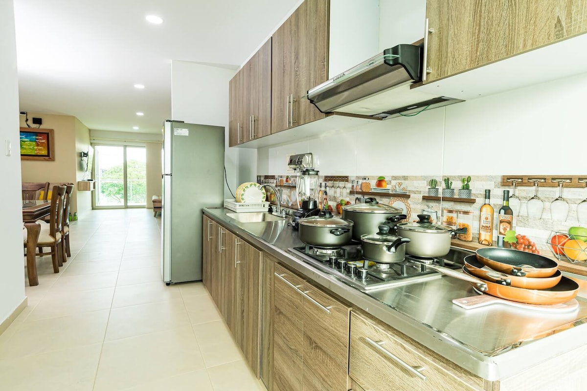 A well-equipped kitchen features sleek wooden cabinetry and a stainless steel stove. Various cookware is neatly arranged on the countertop. A refrigerator stands nearby, with a spacious dining area visible beyond a glass door, allowing natural light to fill the room.