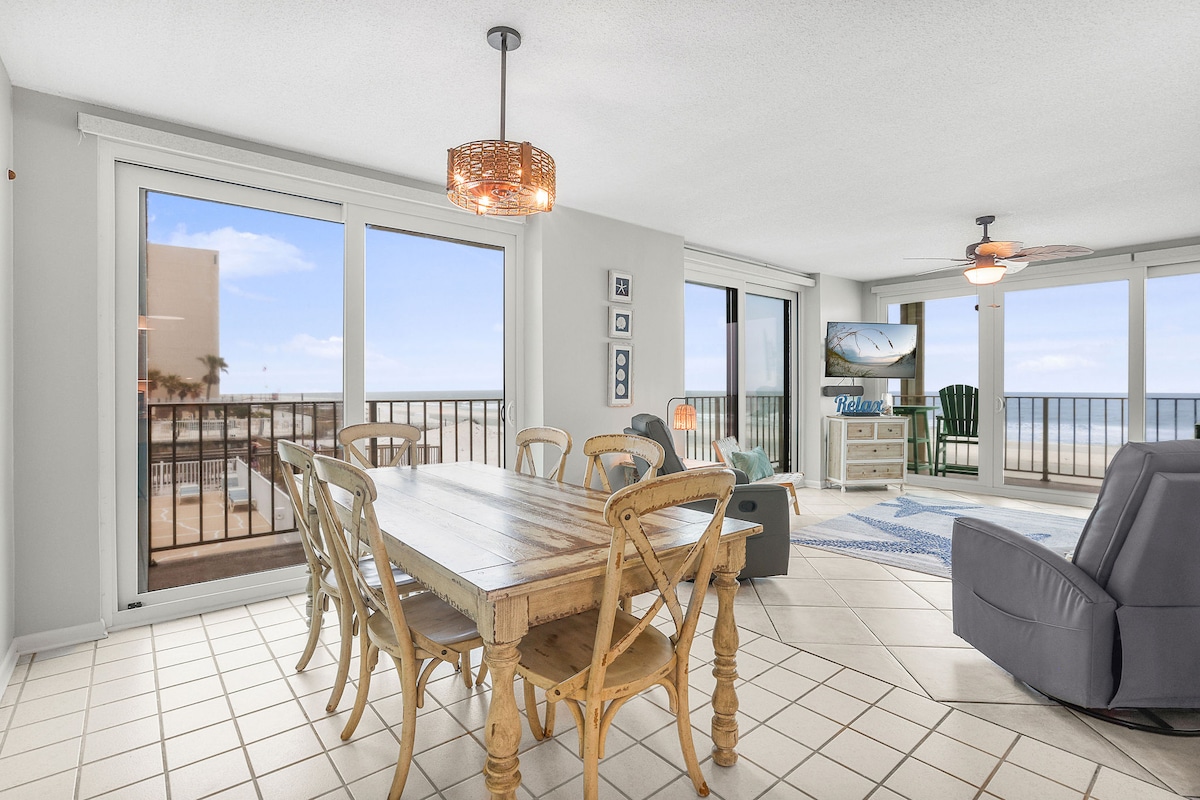 A spacious dining area features a wooden table surrounded by six chairs. Large glass sliding doors provide a view of the beach and allow natural light to fill the space. A TV is visible, positioned for easy viewing from the adjoining living area.
