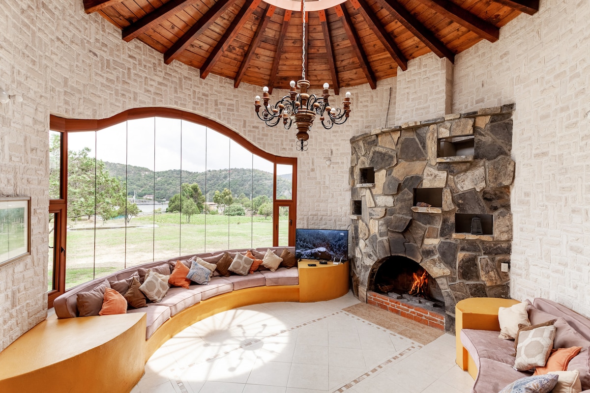 A circular living space features a high wooden ceiling and large windows offering views of the surrounding landscape. Soft seating is arranged along the walls, complemented by a stone fireplace, which adds a cozy touch to the inviting atmosphere.