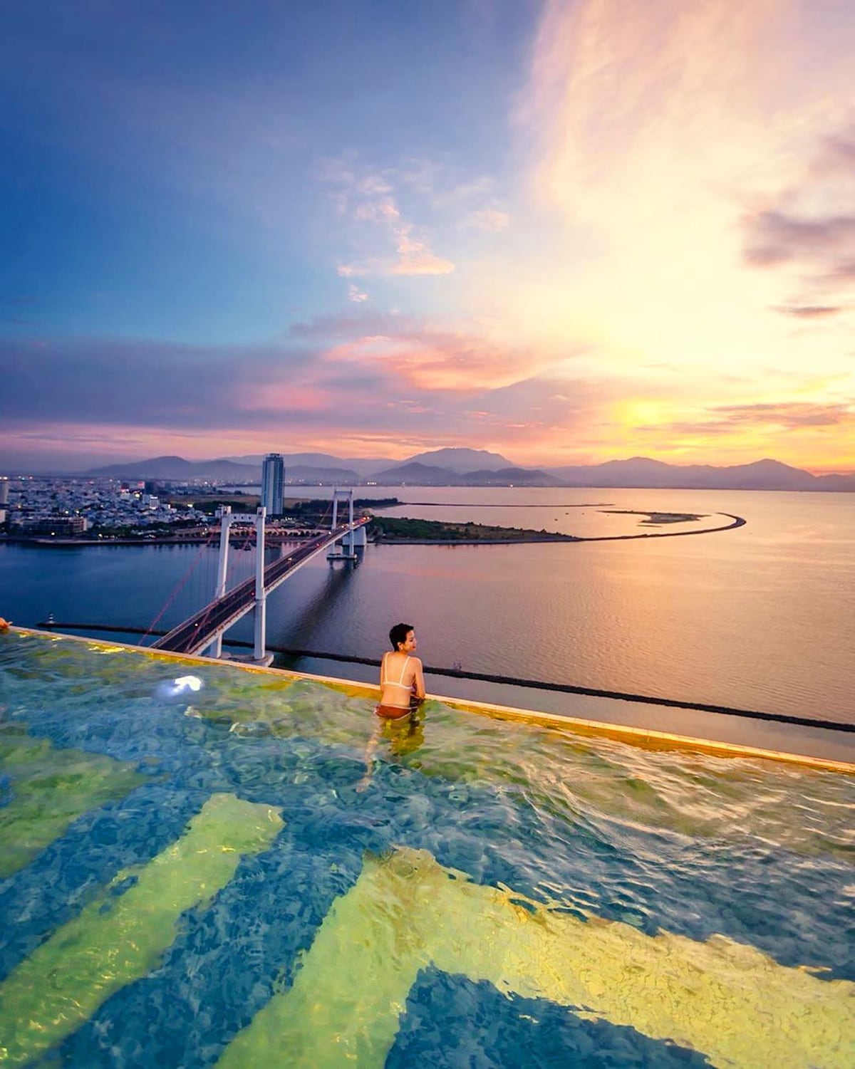 An infinity swimming pool is positioned at a rooftop, overlooking a scenic view of the river and cityscape. The sky transitions into warm hues of orange and purple, reflecting on the water. Gentle waves lap against the pool's edge, creating a serene atmosphere.