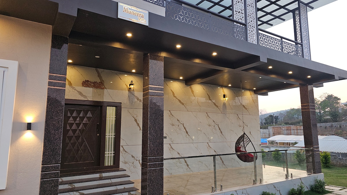 The exterior of the property is characterized by a modern facade featuring elegant marble accents. Bright lights illuminate the entrance, while large glass panels create a seamless transition from the indoors to the outdoors. A decorative wall adds a unique touch to the entrance area.