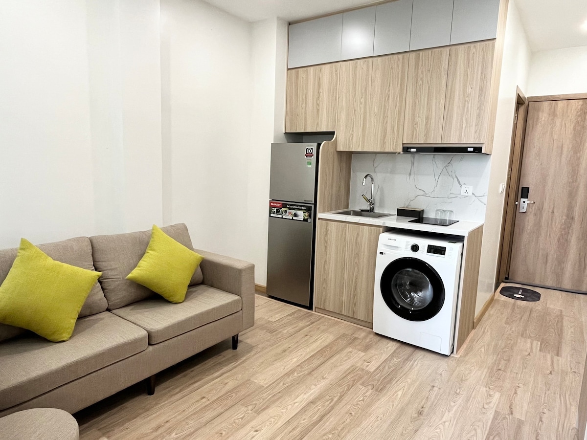 A compact kitchen area features modern appliances, including a refrigerator and washing machine. Light wood cabinetry complements the sleek marble backsplash. A comfortable sofa with two bright yellow accent pillows is positioned nearby, adding a pop of color to the neutral tones of the space.