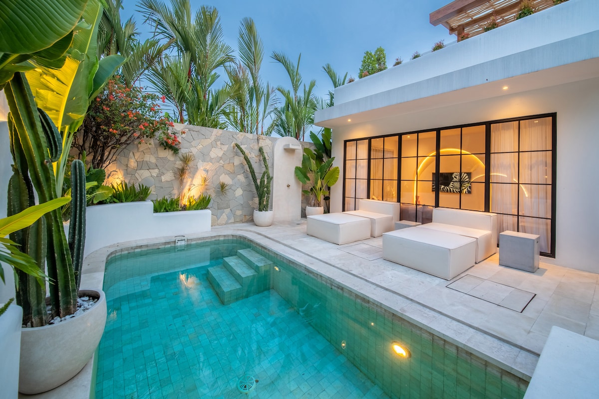 A serene outdoor area features a sparkling pool surrounded by lush greenery and potted plants. White lounge beds are positioned beside the pool, offering comfortable seating. Large glass doors provide access to the indoor space, complementing the tranquil atmosphere.