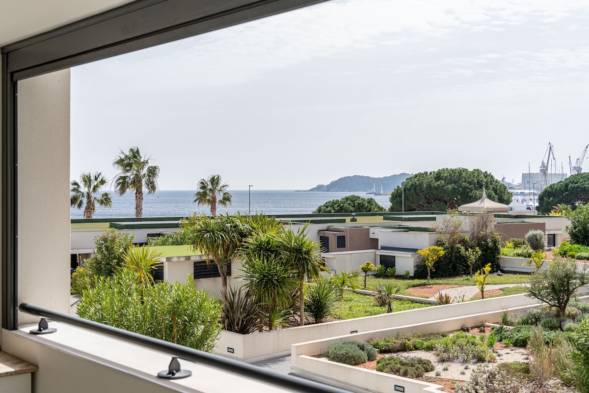 A panoramic view of the Bay of La Ciotat is displayed through a large window. The scene includes lush greenery, palm trees, and neatly arranged gardens below, while the expanse of the sea and distant hills create a peaceful backdrop.