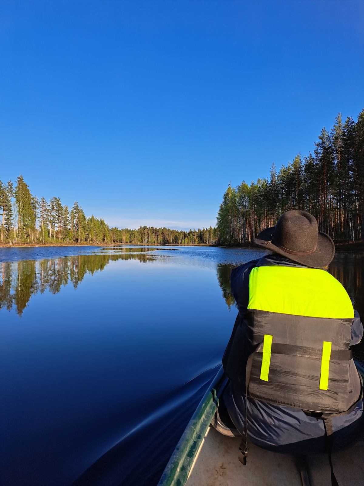 A calm lake is surrounded by tall trees, reflecting the bright blue sky and the greenery. A person in a life jacket sits in a canoe, with their back facing the camera, enjoying the serene landscape.