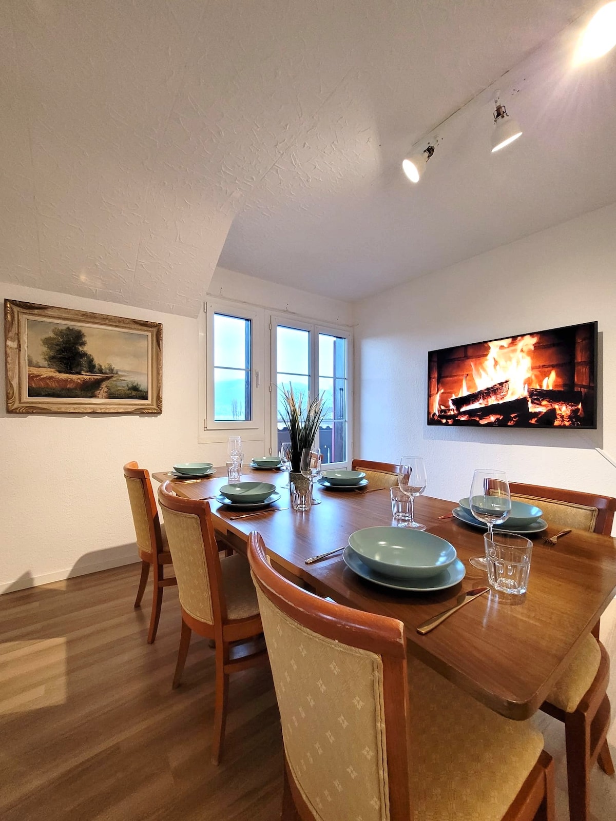 A dining area is displayed, featuring a wooden table set for six with green plates and glassware. A cozy fireplace is mounted on the wall, providing a warm ambiance. Windows allow natural light to enter, highlighting the elegant artwork adjacent to the table.