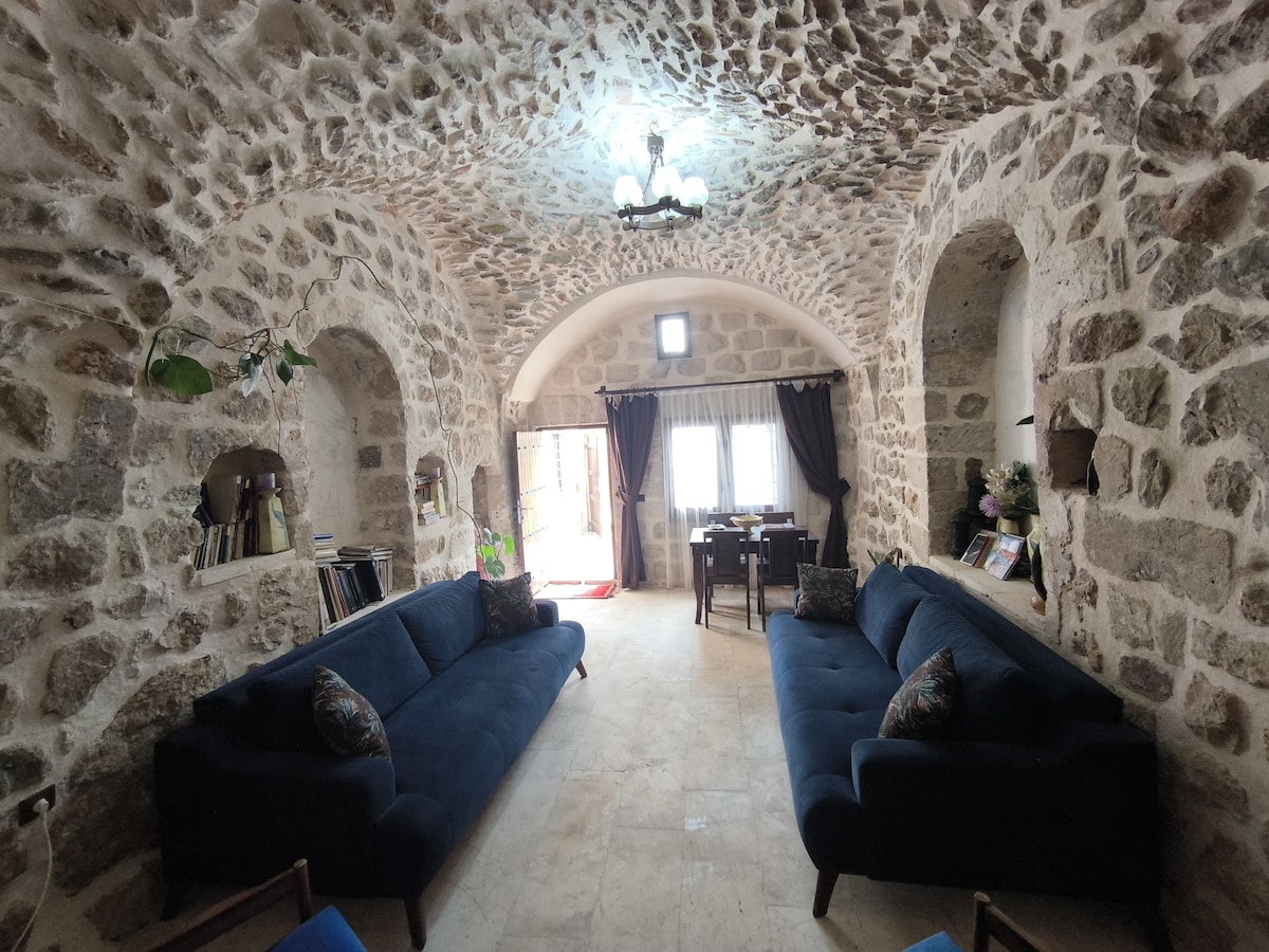 A spacious living area showcases arched stone walls with a textured finish. Two dark blue sofas are placed across from each other, inviting relaxation. Natural light enters through a doorway leading to a dining space, with window coverings enhancing the ambiance.