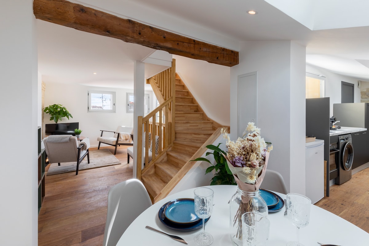 An open-plan living area features a dining table set with blue plates and glassware. A staircase made of light wood extends to an upper level, while a cozy seating arrangement is visible in the background. Natural light enhances the spacious feel of this inviting space.