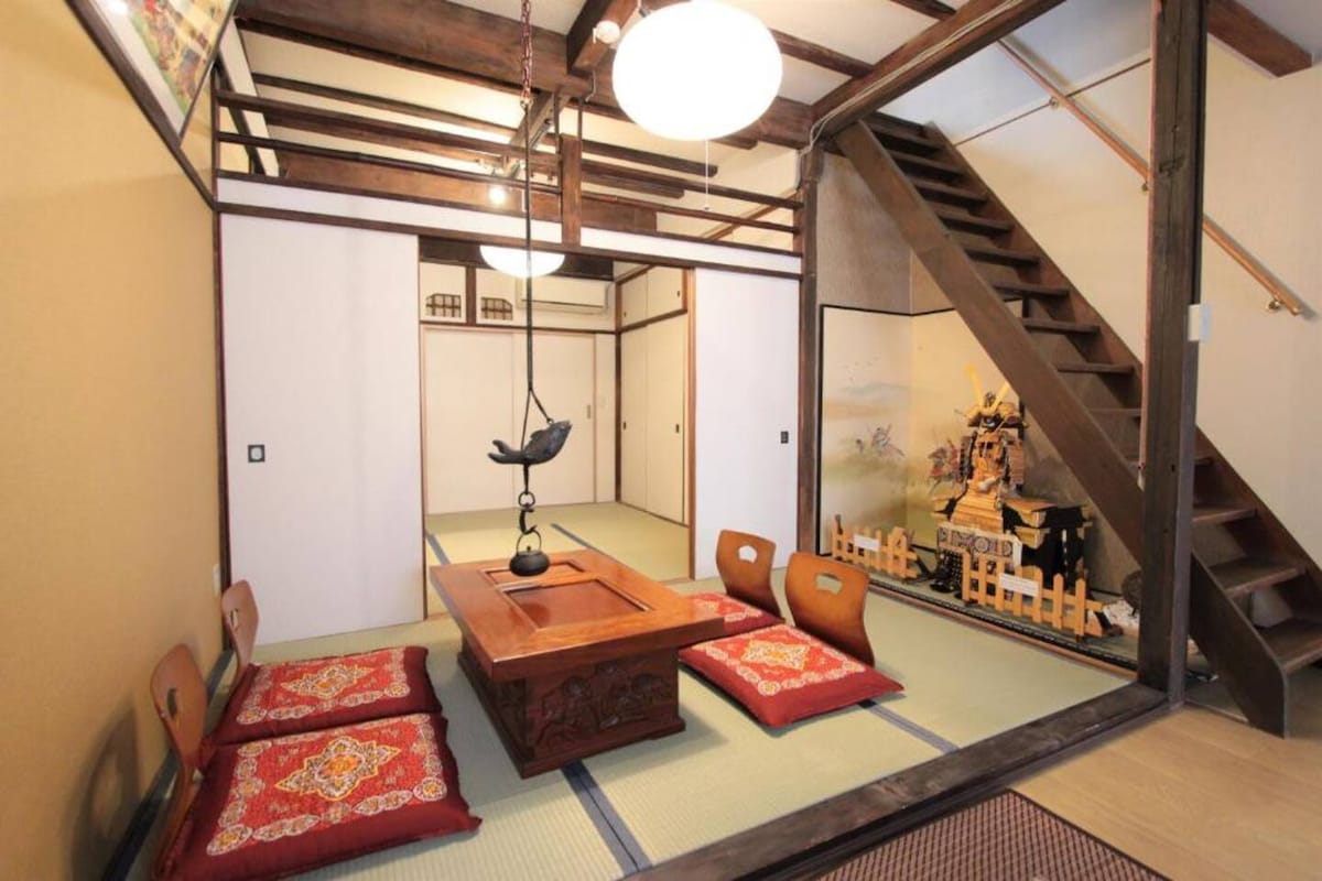 A traditional sitting area features a low wooden table surrounded by floor cushions. The space is framed by tatami mats and offers a view of a staircase leading to the upper level. Natural light filters through overhead fixtures, enhancing the serene ambiance.