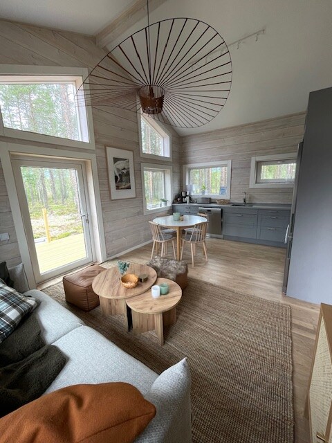 An open living area features large windows allowing for natural light, surrounded by trees. A cozy sofa includes decorative pillows. A round dining table is accompanied by two chairs. The kitchen is equipped with a modern design, integrating seamlessly with the living space.