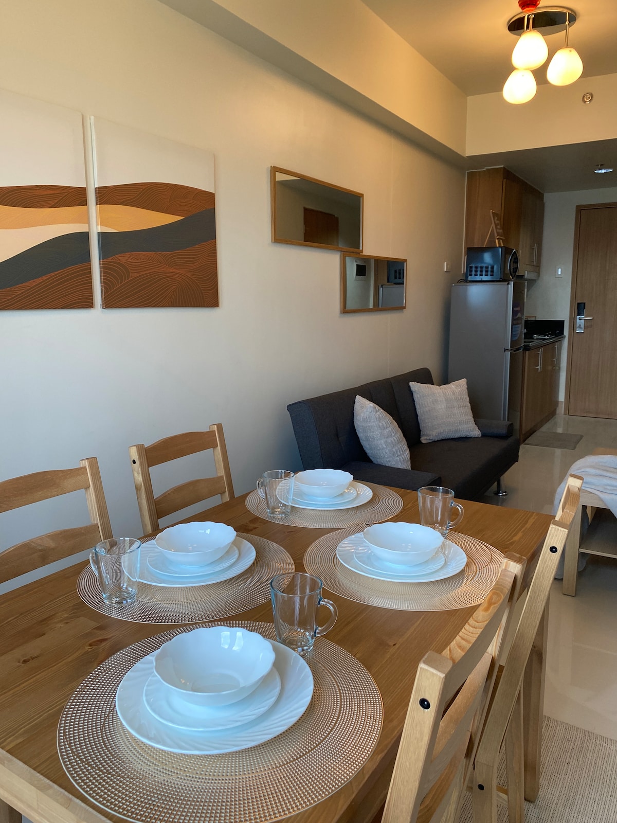 A dining area is featured with a wooden table set for four, including dinnerware and glassware arranged neatly on woven placemats. A comfortable sofa with decorative pillows is positioned nearby, while a modern kitchen is visible in the background.