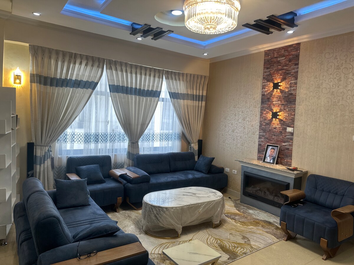 A spacious living area is showcased, featuring large windows adorned with sheer and patterned curtains. Comfortable seating is arranged around a round coffee table, with a fireplace providing a focal point. Subtle lighting accents the ceiling, enhancing the interior design.