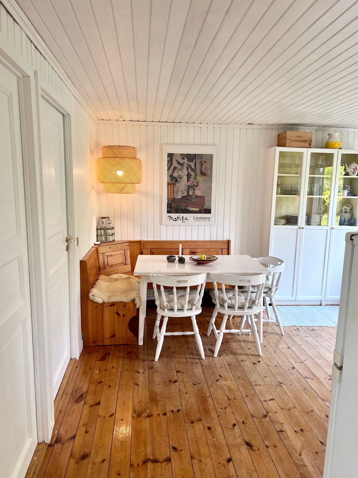 A dining area features a white table surrounded by wooden chairs, complemented by a built-in bench. Natural light illuminates the space, enhancing the wood-paneled walls and floor. A light fixture hangs above, adding a cozy touch to the inviting atmosphere.