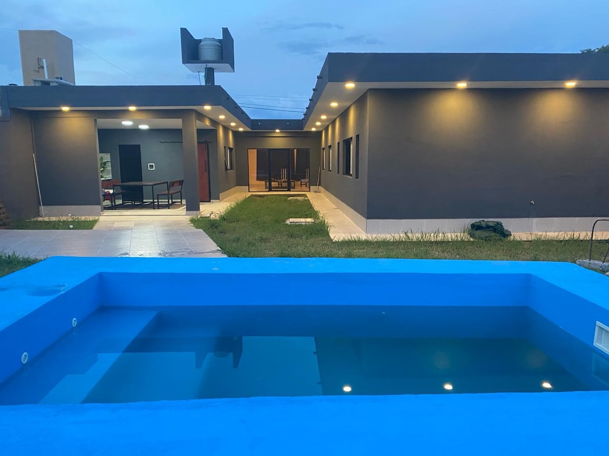 A vibrant blue pool is set in a landscaped area, reflecting the evening sky. The modern single-story house features a patio with seating visible through large windows, surrounded by well-maintained grass. Soft outdoor lighting enhances the structure's sleek exterior.