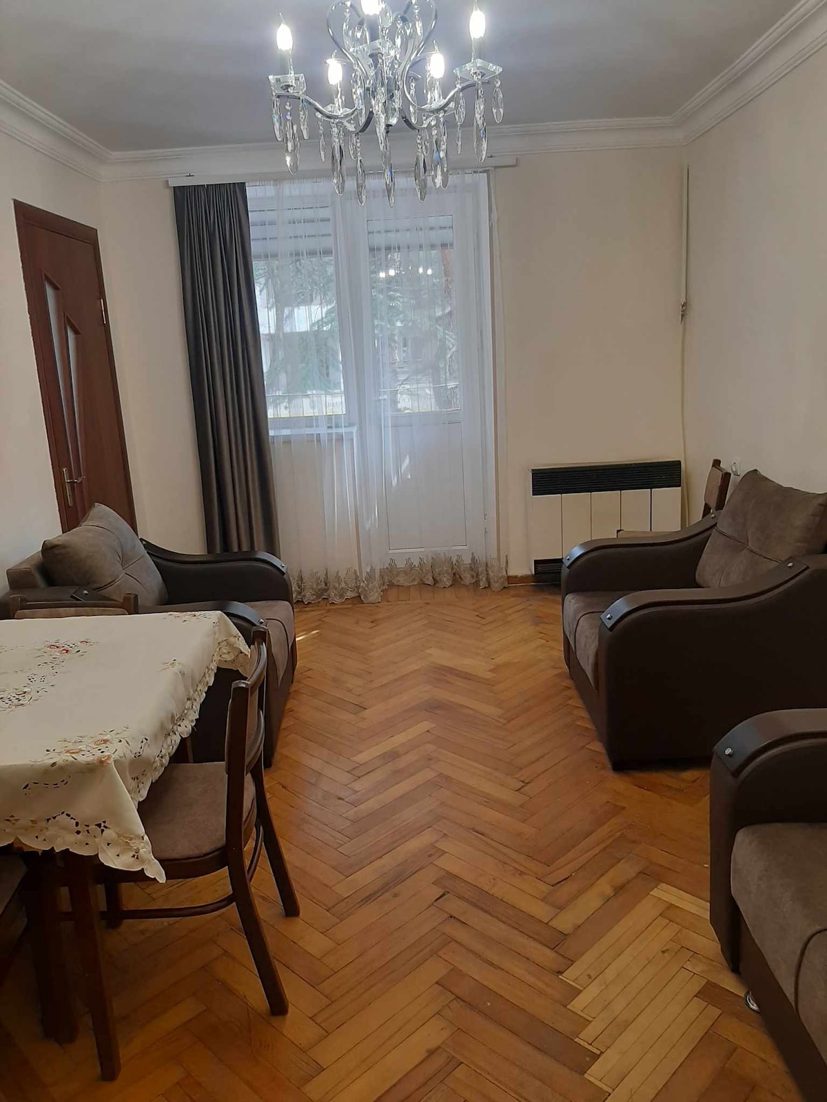 A comfortable living area features a dining table with a decorative tablecloth and four chairs. Two armchairs are positioned nearby. Natural light enters through large windows adorned with sheer curtains, illuminating the parquet flooring and creating a cozy environment.