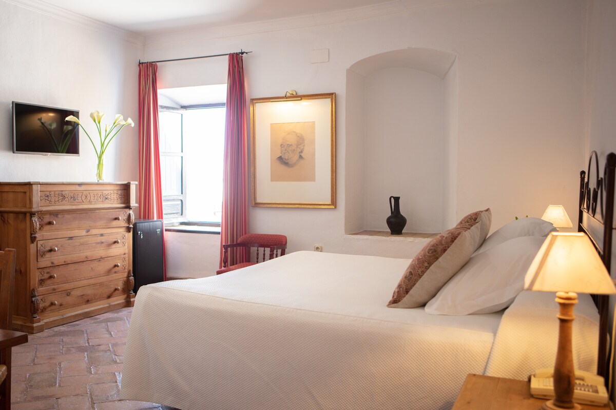 A spacious room is illuminated by natural light from a window, featuring a comfortable bed with decorative pillows. A wooden dresser and a small refrigerator are present, along with a television mounted on the wall. Elegant curtains frame the window, enhancing the room's tranquil ambiance.