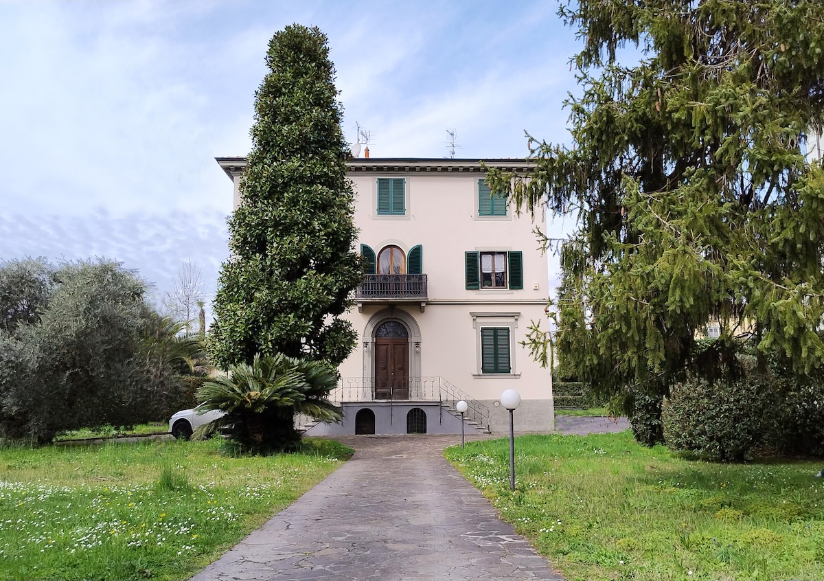 A historic villa is viewed from a pathway lined with greenery. The structure features large windows and a decorative balcony. Surrounding the villa, landscaped gardens with various trees create a serene environment. The entrance is clearly marked by stylish outdoor lighting.