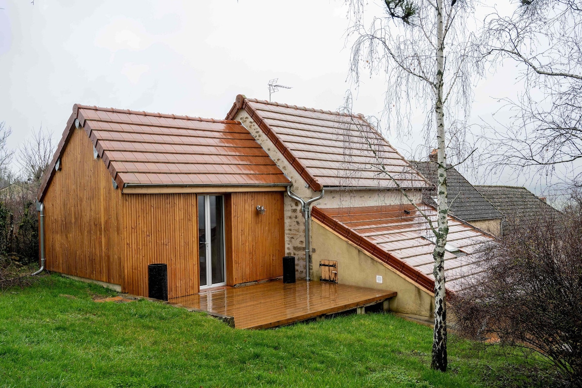 A wooden-clad structure with large glass doors overlooks a grassy area. The roof features terracotta tiles, and the building is set against a backdrop of a cloudy sky. A birch tree stands nearby, complementing the natural surroundings.