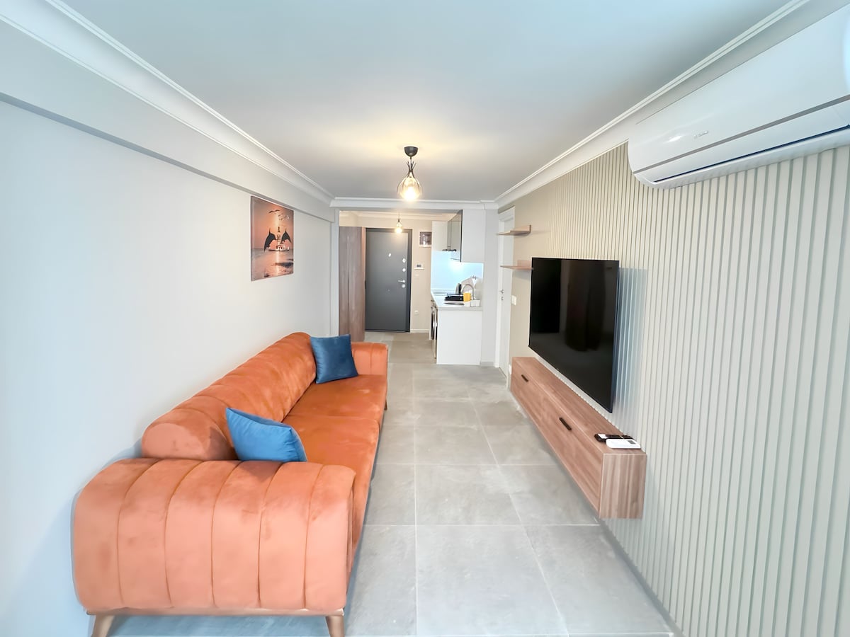 A modern living area is showcased with a vibrant orange sofa adorned with blue cushions. A wall-mounted TV is positioned across from the seating, while a light fixture hangs from the ceiling, illuminating the space. A glimpse of the kitchen is visible in the background.