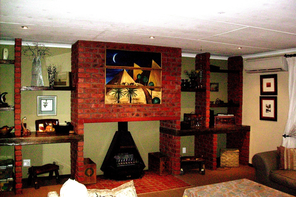 The living area features exposed brick walls with built-in shelves displaying decorative items. A large painting depicting desert landscapes is prominently displayed. A cozy seating arrangement is set around a central fireplace, contributing to a warm and homely environment.