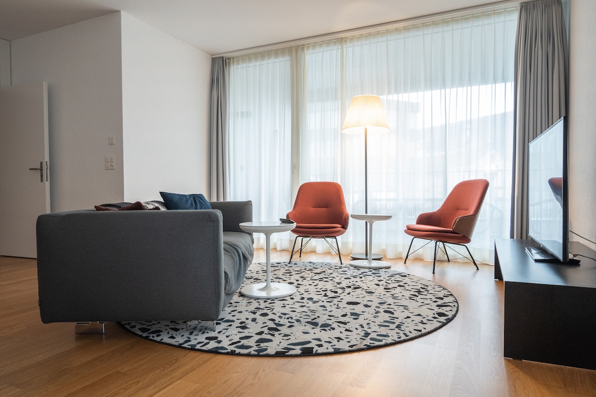 A modern living area features a sleek gray sofa, complemented by two orange armchairs and a circular white coffee table. A neutral area rug enhances the warmth of the wooden flooring. Natural light filters through sheer curtains, providing a bright and airy atmosphere.