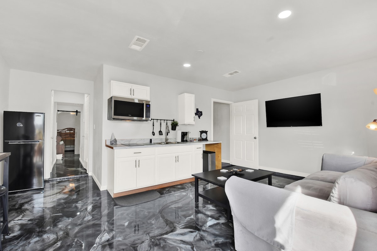 The open living area combines a modern kitchen with essential appliances, including a fridge and microwave. A comfortable seating arrangement is positioned in front of a wall-mounted television, while the light-toned walls enhance the space's brightness. The polished flooring contributes to the contemporary aesthetic.